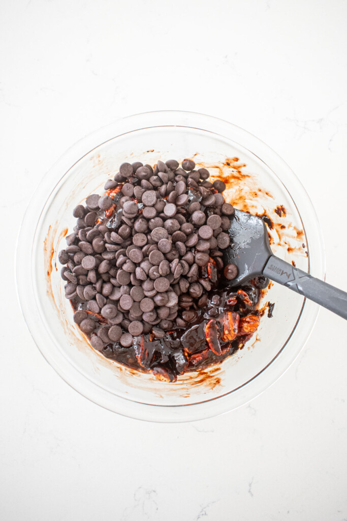 chocolate bourbon pecan pie filling with pecans being stirred in a glass mixing bowl on a white marble counter.