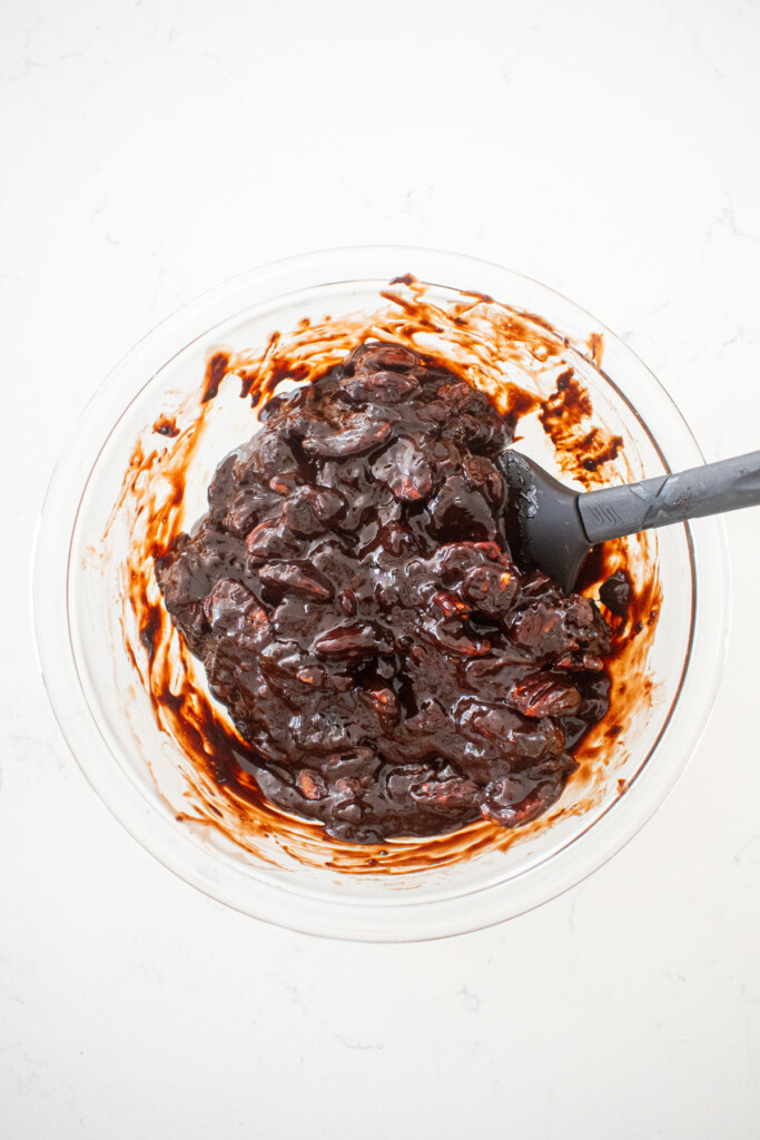 chocolate bourbon pecan pie filling with pecans being stirred in a glass mixing bowl on a white marble counter.