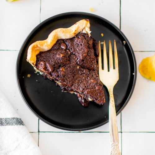 slice of chocolate bourbon pecan pie on a black plate on a white tile countertop.