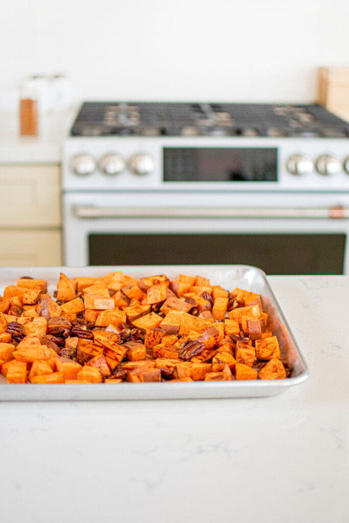 cinnamon sweet potatoes and pecans on a baking sheet on a white marble kitchen countertop.