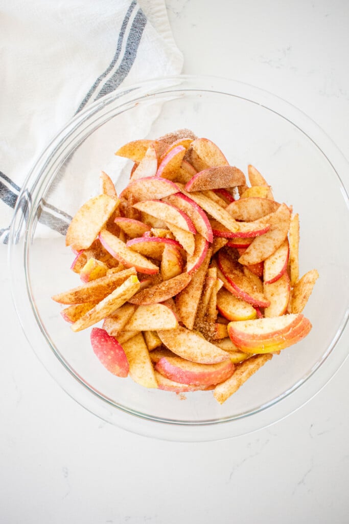 apples mixed together with sugar in a glass mixing bowl on a marble counter.