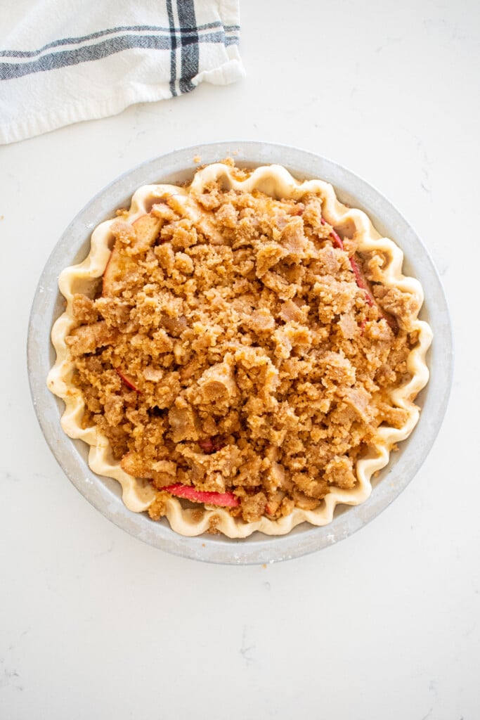 crumb topping put on top of an apple pie in a pie pan on a marble counter.