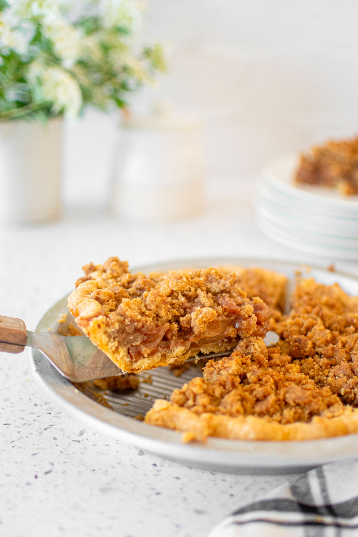 old fashioned dutch apple pie slice being lifted from a pie pan on a white marble counter.