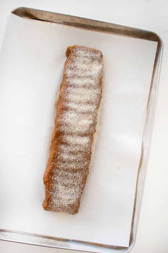 gingerbread biscotti cookie dough log sprinkled with cane sugar on a baking sheet on a marble counter.