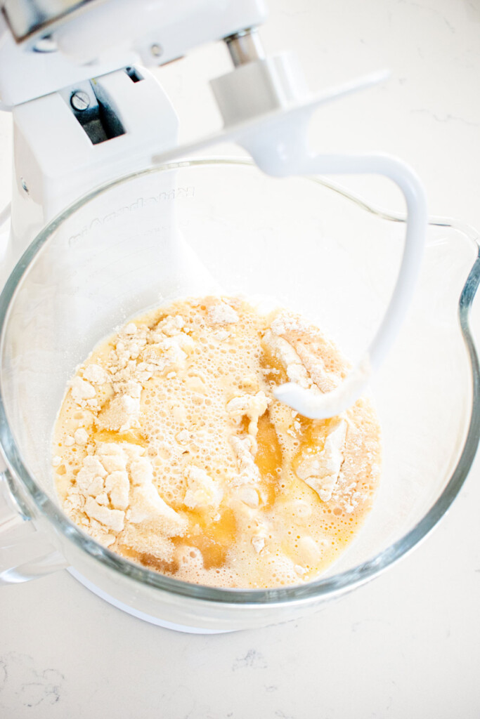 ingredients for honey butter dinner rolls in a glass mixing bowl on a white marble counter.