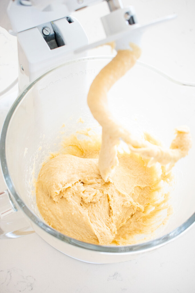 honey butter dinner rolls dough in a glass mixing bowl with a dough hook on a white marble counter.