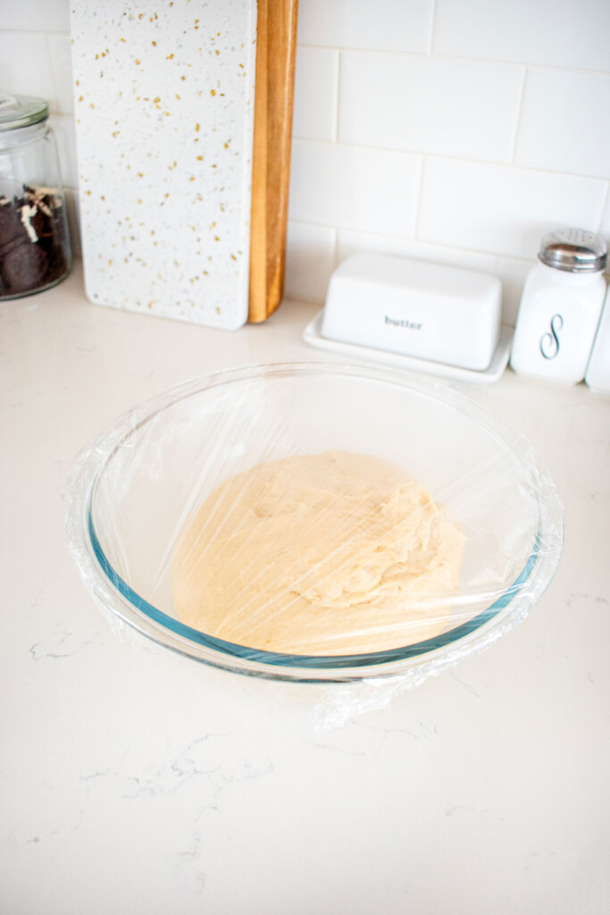 honey butter dinner rolls dough in a glass mixing bowl on a white marble counter.