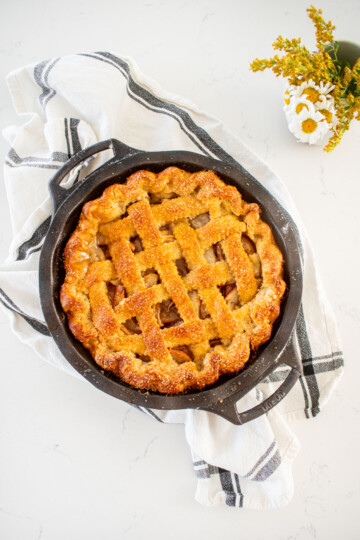 golden brown orange blossom honey apple pie with a lattice top in a cast iron pie pan on a marble counter with a small vase of flowers.