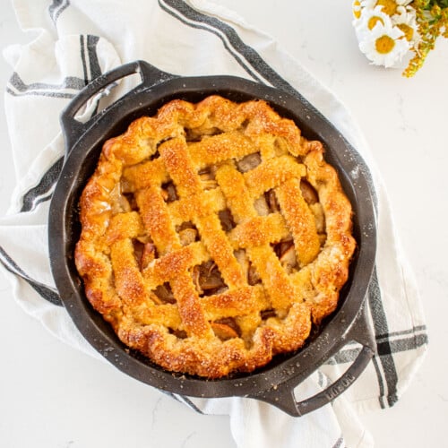 golden brown orange blossom honey apple pie with a lattice top in a cast iron pie pan on a marble counter with a small vase of flowers.