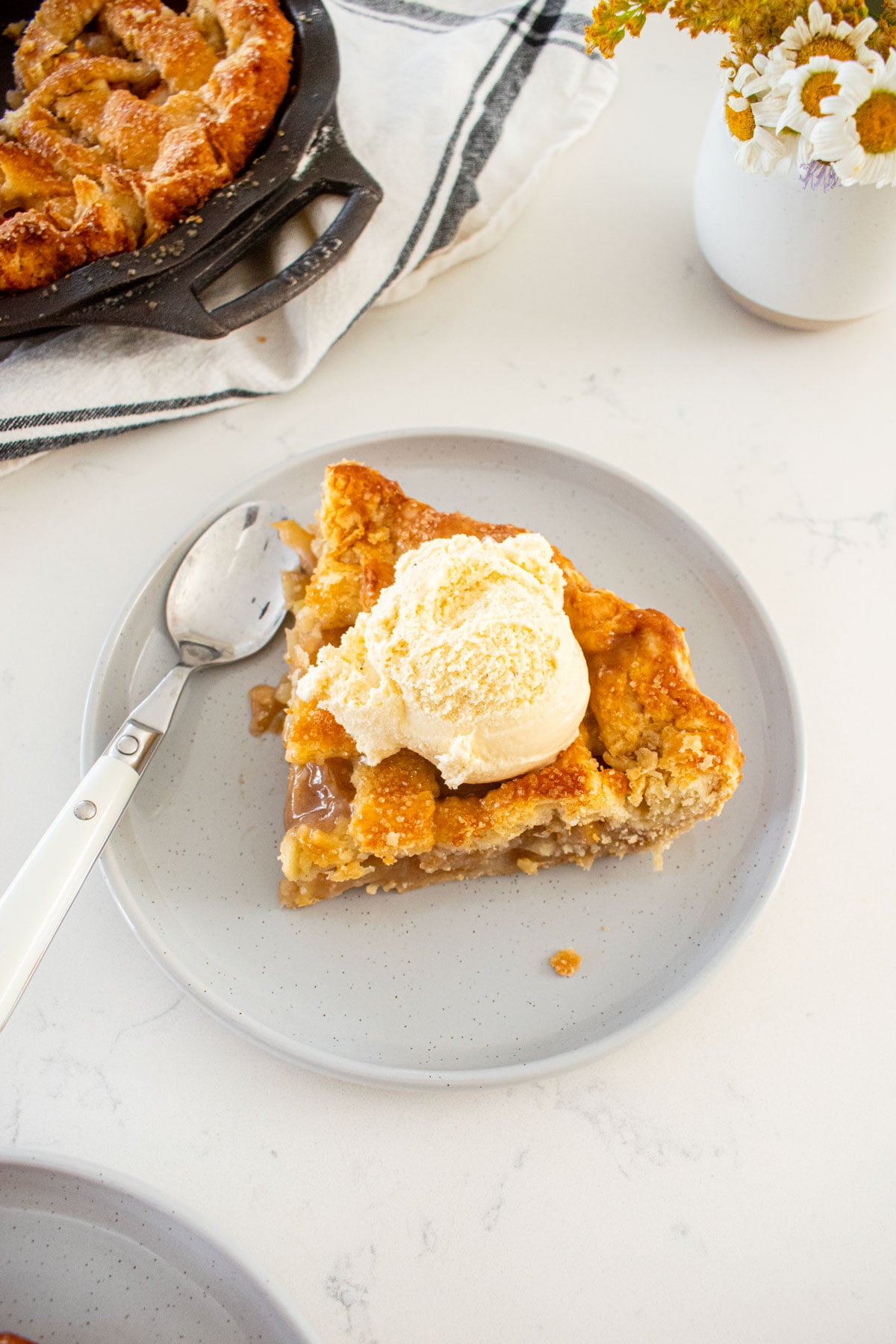slice of orange blossom honey apple pie on a plate with a scoop of vanilla ice cream on a marble counter.