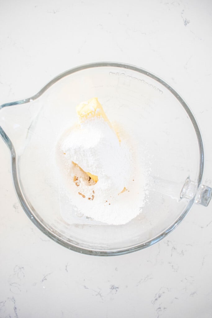 butter, powdered sugar and vanilla extract in a glass mixing bowl on a white marble counter.