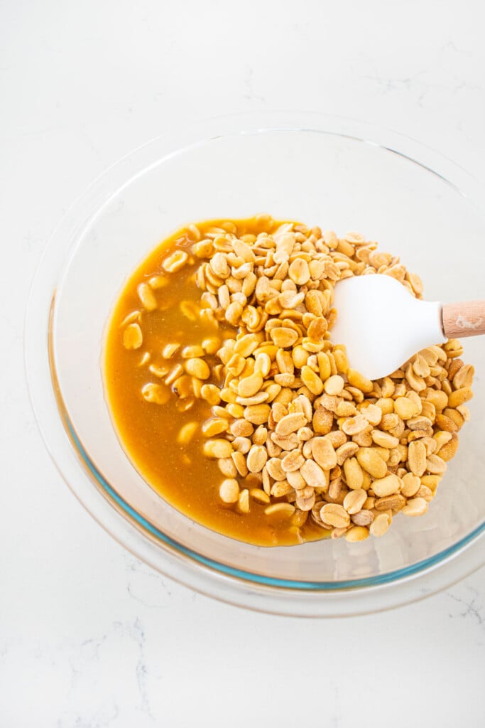 peanuts and sugar mixture being mixed in a glass mixing bowl on a white marble counter.