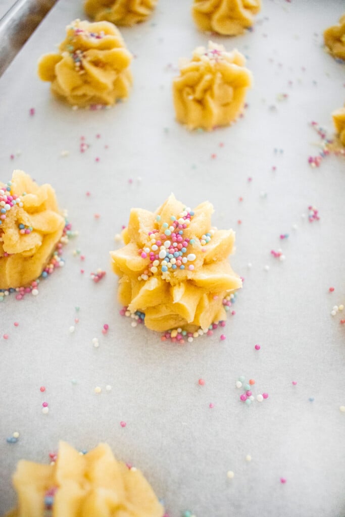 piped eggless spritz cookie dough with sprinkles on top on a baking sheet on a marble counter.