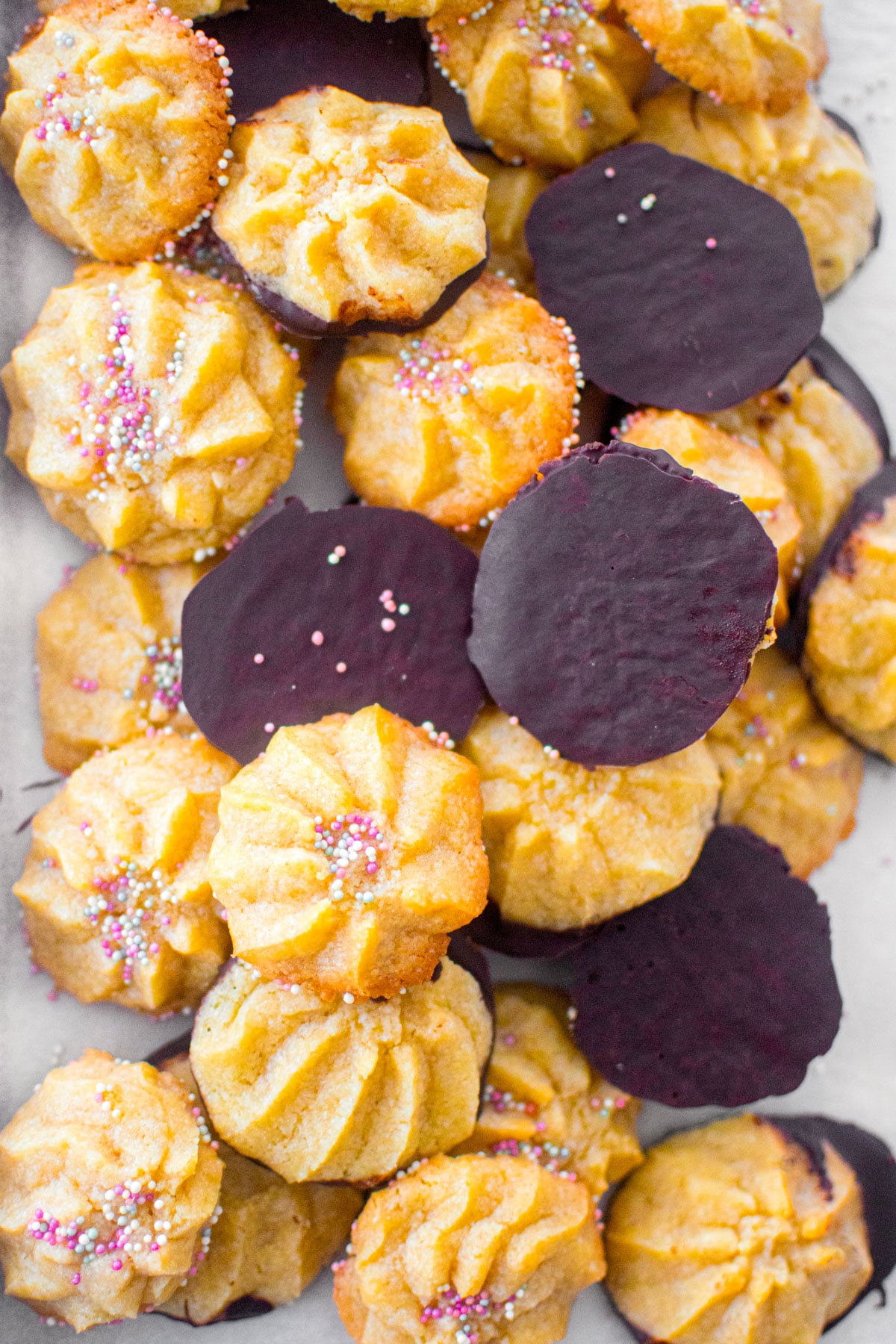 eggless spritz cookies half dipped in chocolate and some with sprinkles on a baking pan on a marble counter.