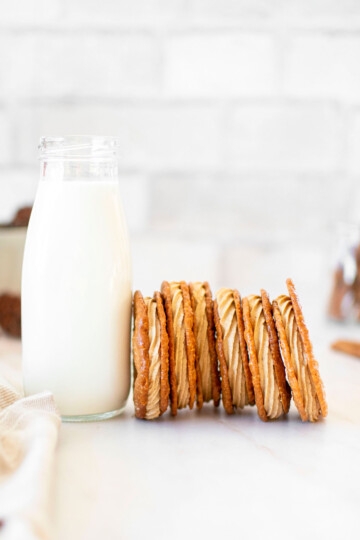 gingerbread florentine sandwich cookies leaning up against a glass milk bottle on a white marble counter.
