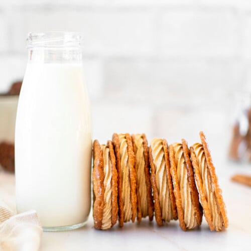 gingerbread florentine sandwich cookies leaning up against a glass milk bottle on a white marble counter.