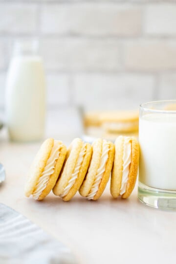 homemade vanilla oreo cookies leaning up against a glass of milk on a marble countertop.