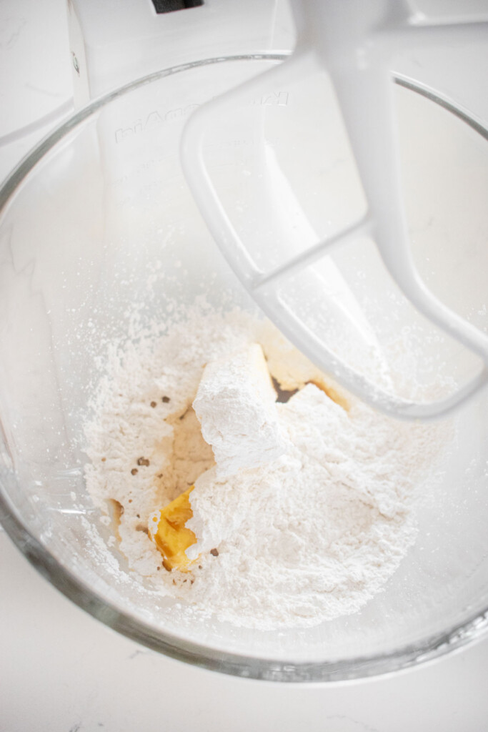 butter, powdered sugar and vanilla in a glass mixing bowl on a white marble countertop.