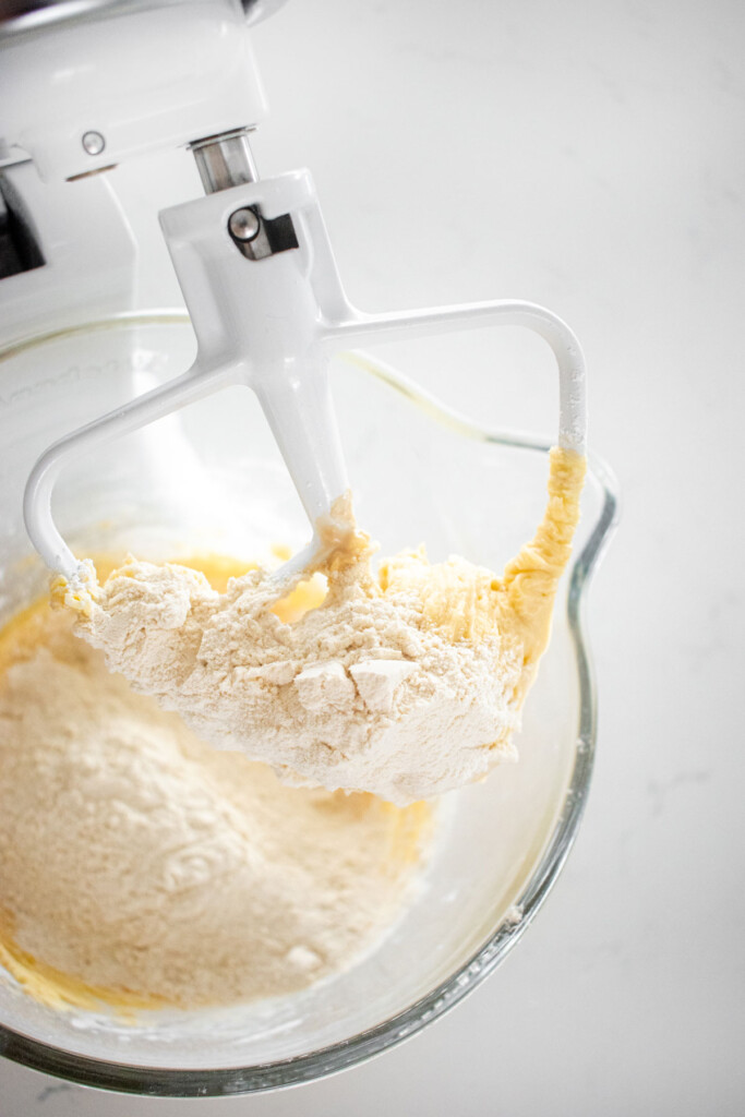 flour added into butter mixture for vanilla shortbreads in a glass mixing bowl on a white marble countertop.