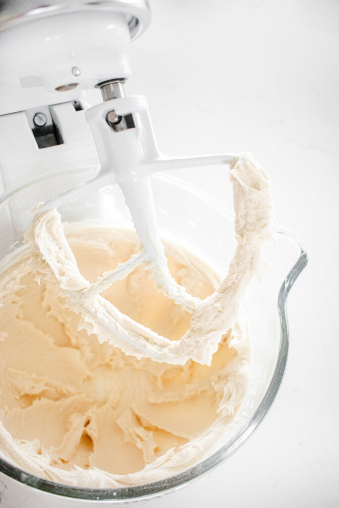 vanilla buttercream mixed in a stand mixer in a glass bowl on a white marble countertop.
