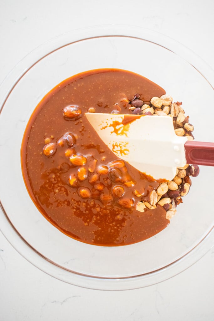 caramel being added to a glass bowl with peanuts on a white marble countertop.