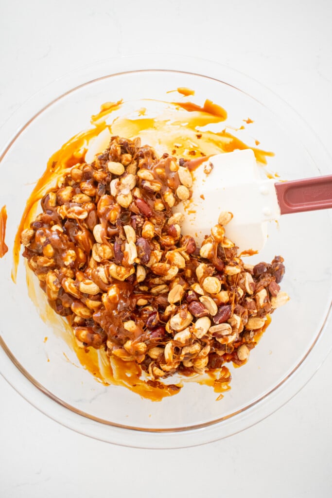 caramel being stirred with peanuts in a glass bowl on a white marble countertop.