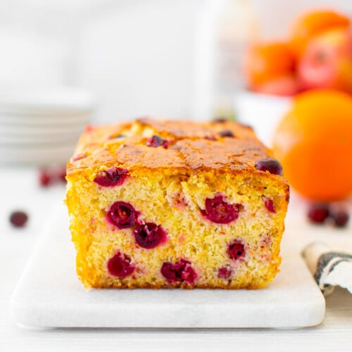 cranberry orange apple bread on a white marble board with plates fresh oranges and apples in the background.