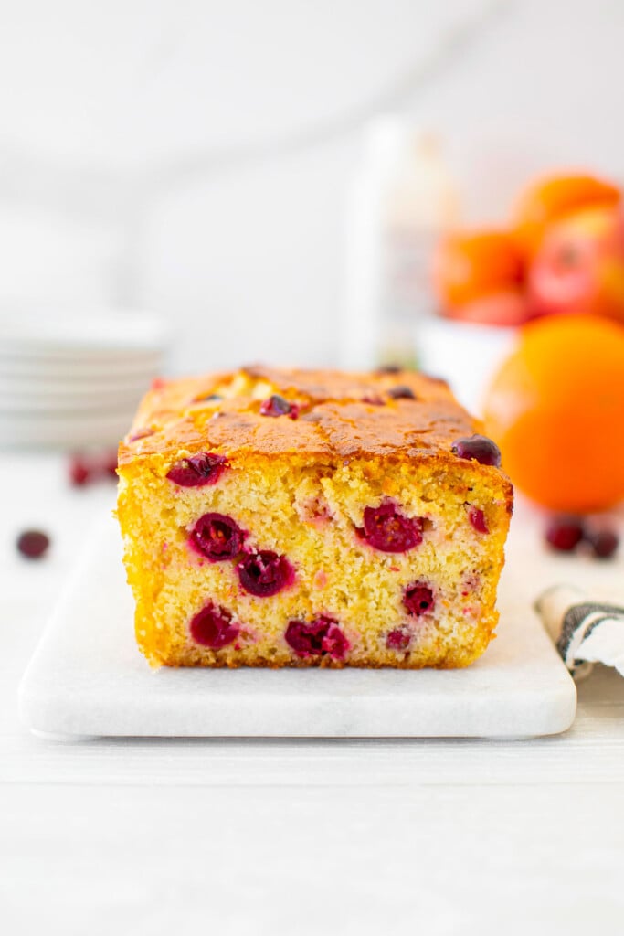 cranberry orange apple bread on a white marble board with plates fresh oranges and apples in the background.