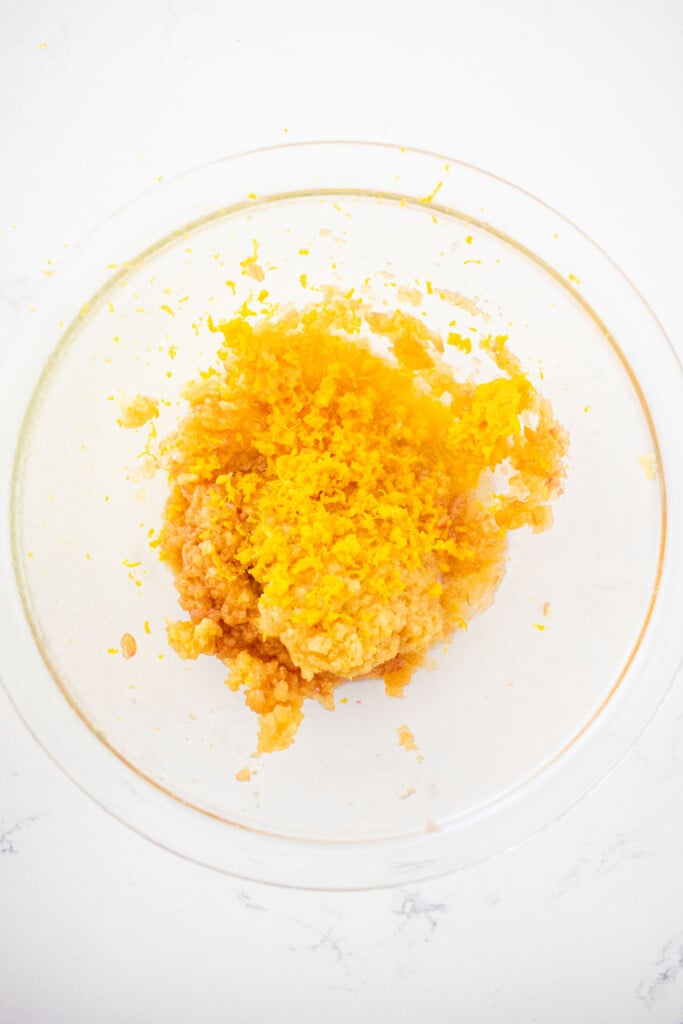 grated apple and orange zest in a glass mixing bowl on a white marble counter.