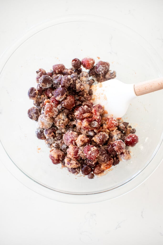 glass bowl with cherries being mixed with a spatula into pie filling on a white marble counter.