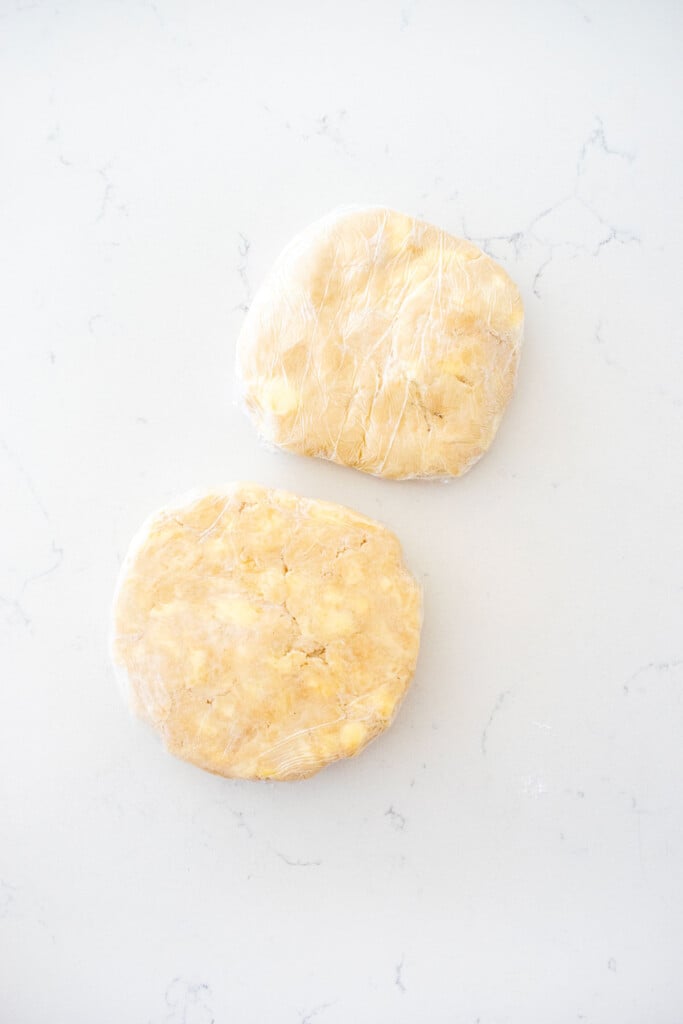 two pie crusts shaped into disks wrapped in plastic wrap on a white marble counter.