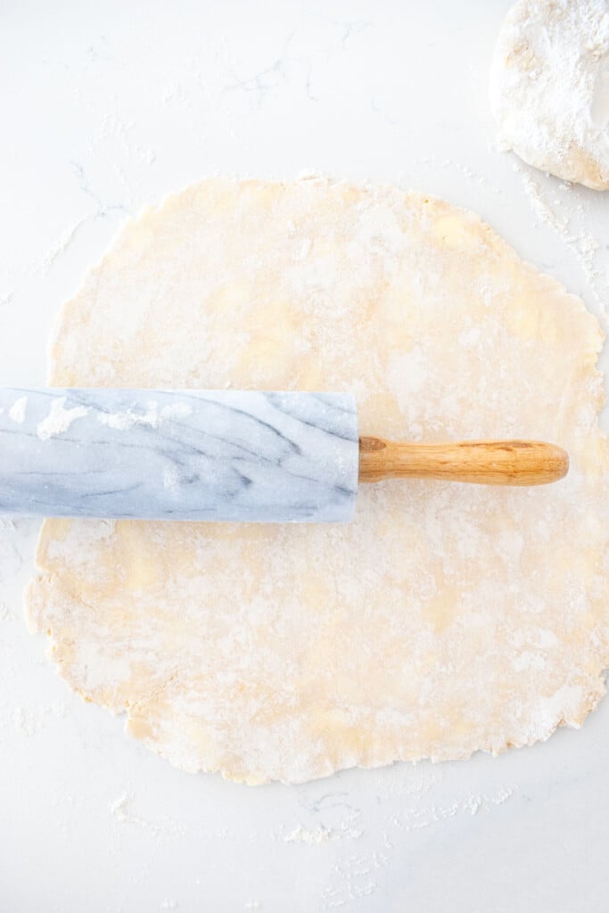 pie crust being rolled out with a marble rolling pin on a white counter.
