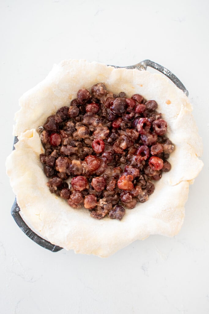 cast iron pie pan with pie crust and cherry pie filling on a white marble counter.