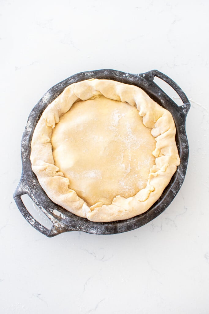 cherry pie being assembled in a black cast iron pie pan on a white marble counter.