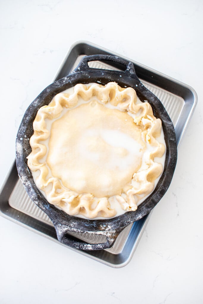 cherry pie with fluted crust and milk brushed on top in a black cast iron pie pan on a white marble counter.