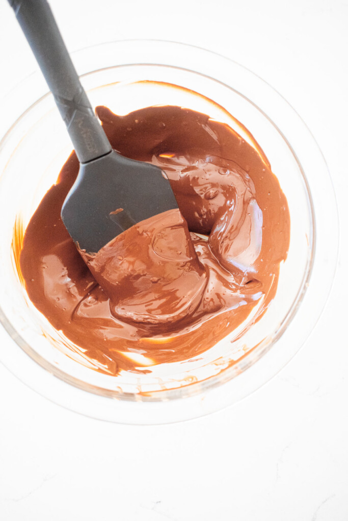 melted chocolate in a glass mixing bowl with a black spatula on a marble counter.