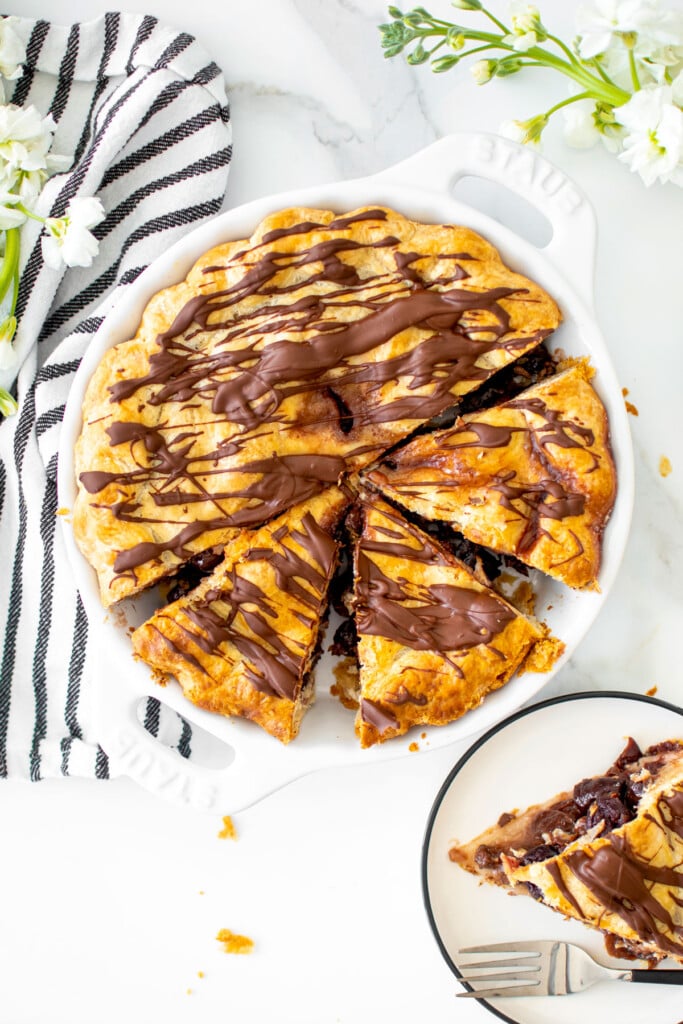 cherry chocolate chip pie cut into slices in a white pie dish on a white marble table with fresh flowers.
