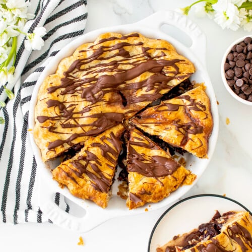cherry chocolate chip pie cut into slices in a white pie dish on a white marble table with fresh flowers.