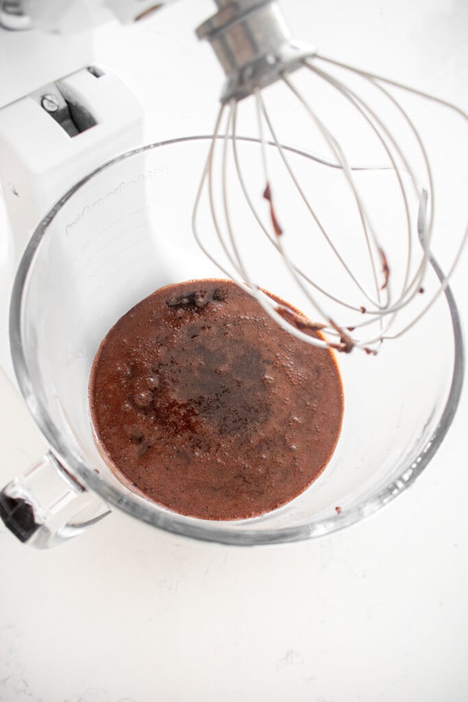 ingredients for homemade chocolate marshmallows in a glass stand mixing bowl with a whisk on a white marble counter.