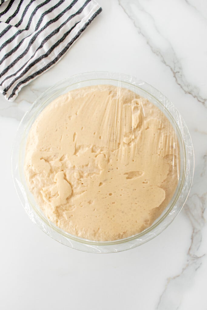 glass mixing bowl of proofed hamburger bun dough on a white marble counter.