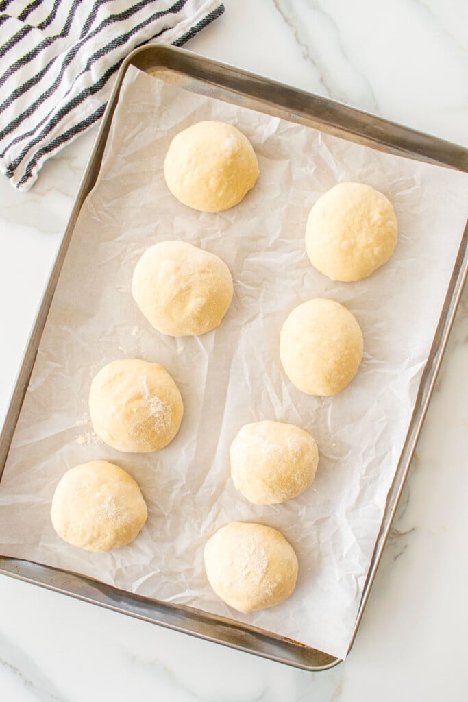 white hamburger bun dough formed into dough balls on a baking sheet on a marble counter.