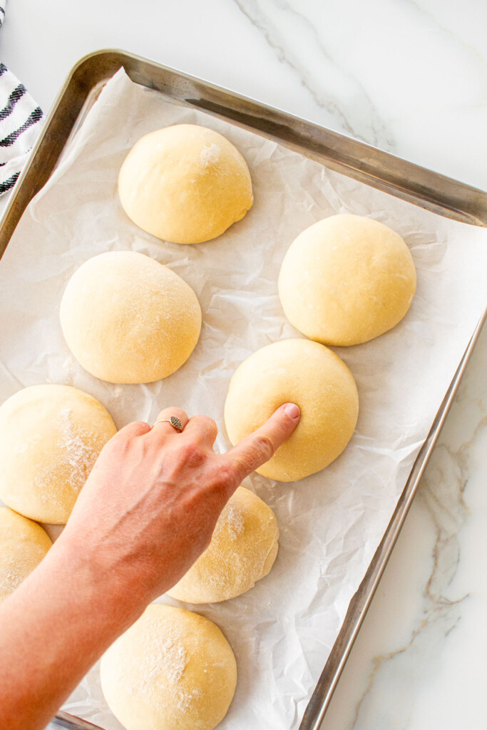 woman pressing finger into hamburger bun dough on a baking sheet to test springy texture.
