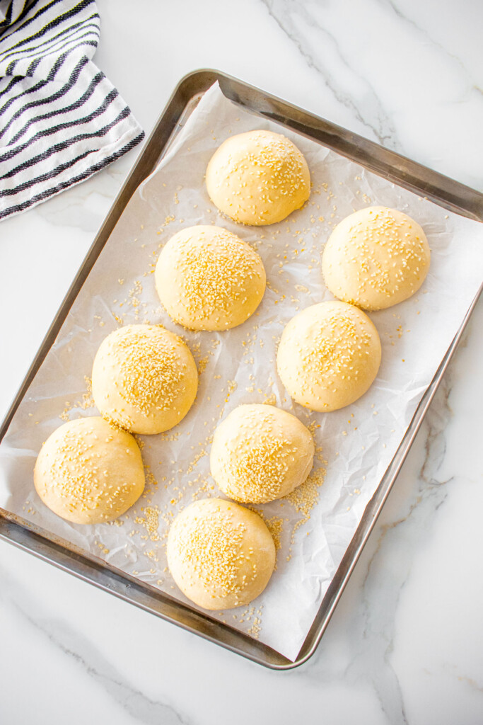 soft white hamburger bun dough balls with sesame seeds on top on a baking sheet on a marble counter.