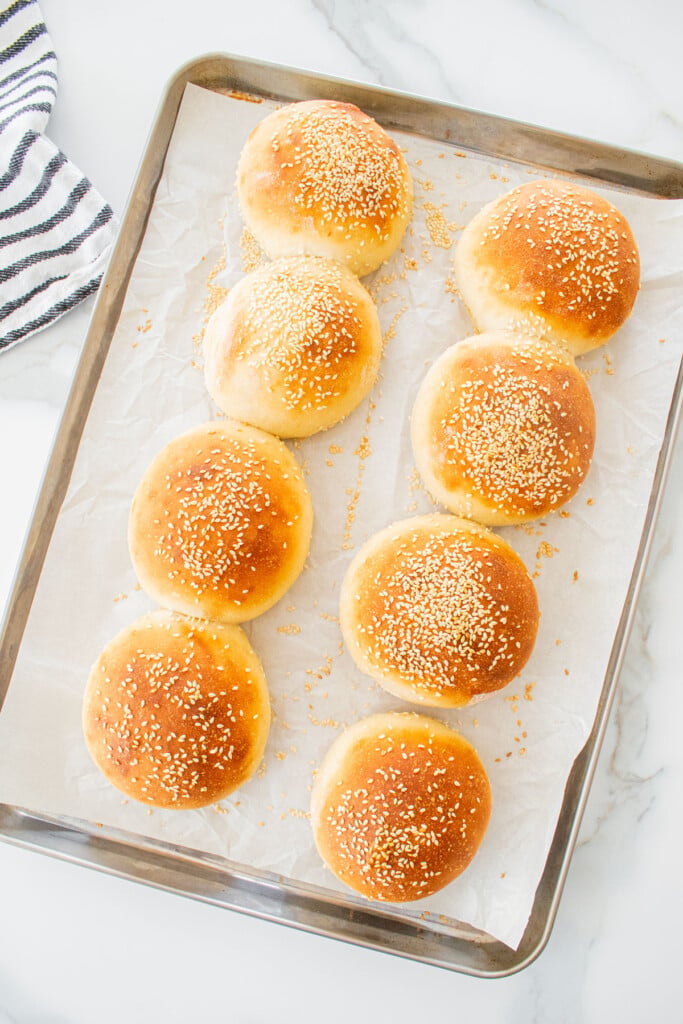 golden brown baked soft white hamburger buns that are golden brown on a baking sheet on a marble counter.