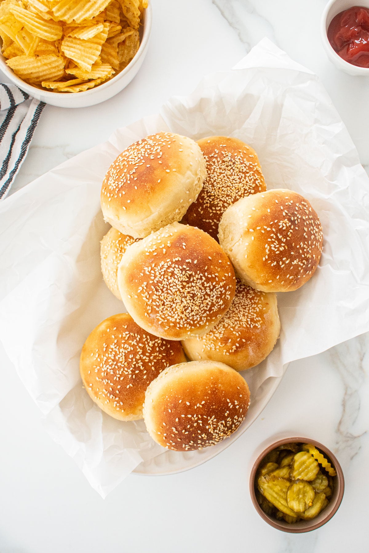 soft white hamburger buns with sesame seeds on a serving platter on a white marble counter with burger toppings in bowls.