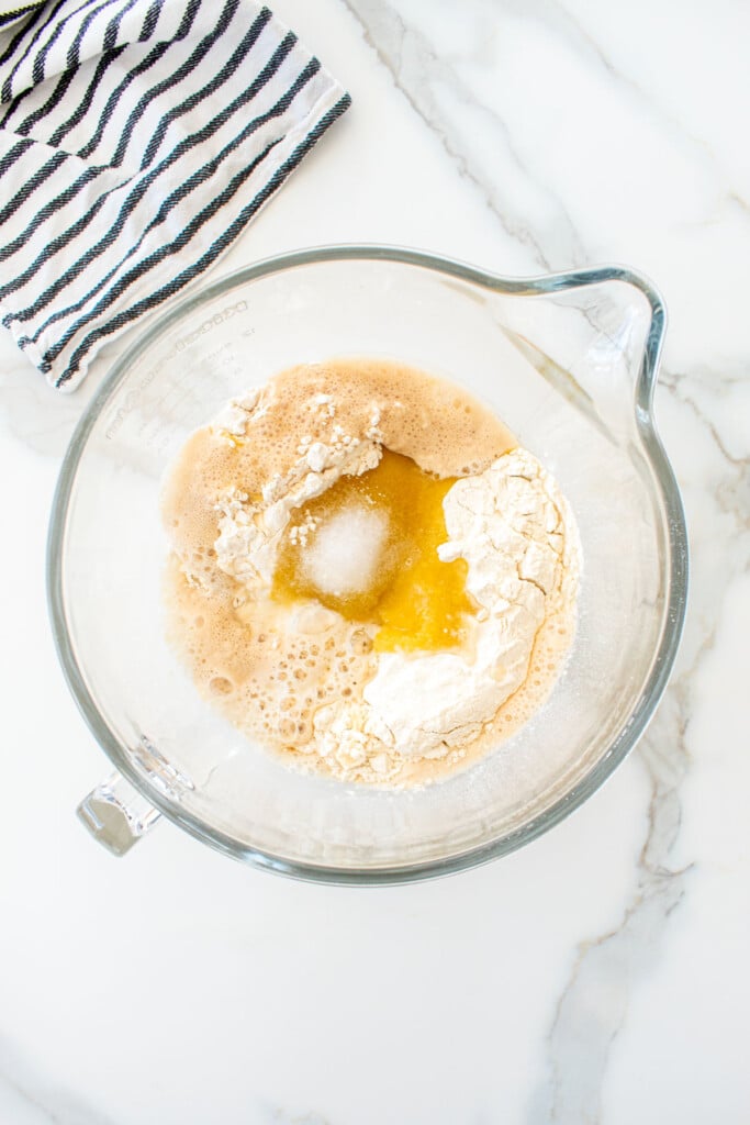 ingredients for soft white hamburger buns in a glass mixing bowl on a white marble counter.