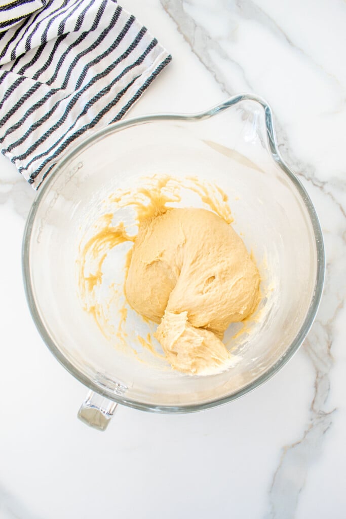 soft white hamburger bun dough in a glass mixing bowl on a white marble counter.