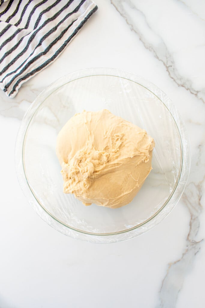 soft white hamburger bun dough in a glass mixing bowl covered with plastic wrap on a white marble counter.