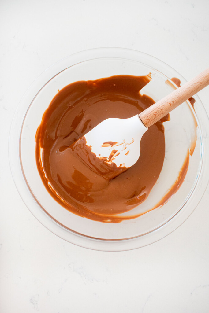 melted milk chocolate in a glass bowl on a white marble counter with a spatula in it.