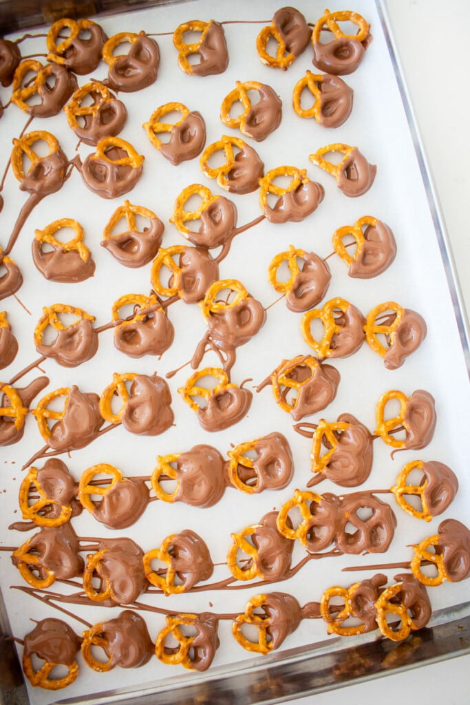 mini pretzel twists dipped in milk chocolate on a baking sheet with parchment paper on a marble counter.